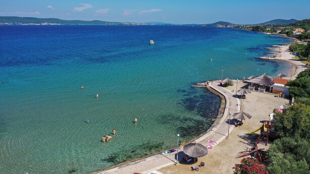 A coastal beach with clear waters, a boat, and a beachside area with umbrellas and seating. - Villa (Dobropoljana)