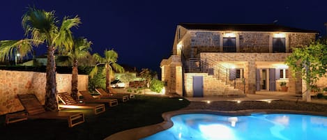 A stone building with a swimming pool, palm trees, and lounge chairs.
