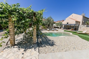 Ein Poolbereich mit Liegestühlen, einer mit Weinreben bewachsenen Pergola und einem Steinhaus mit Ziegeldach.