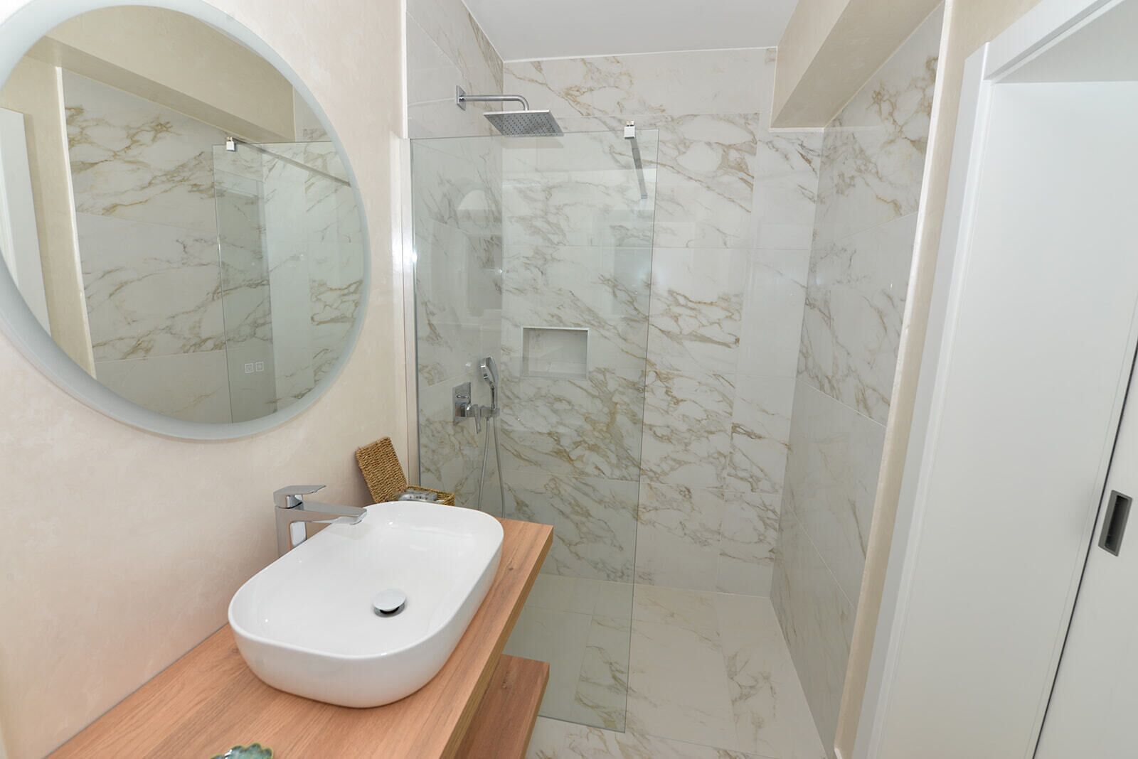 A modern bathroom with a marble shower, a round mirror, and a wooden vanity.