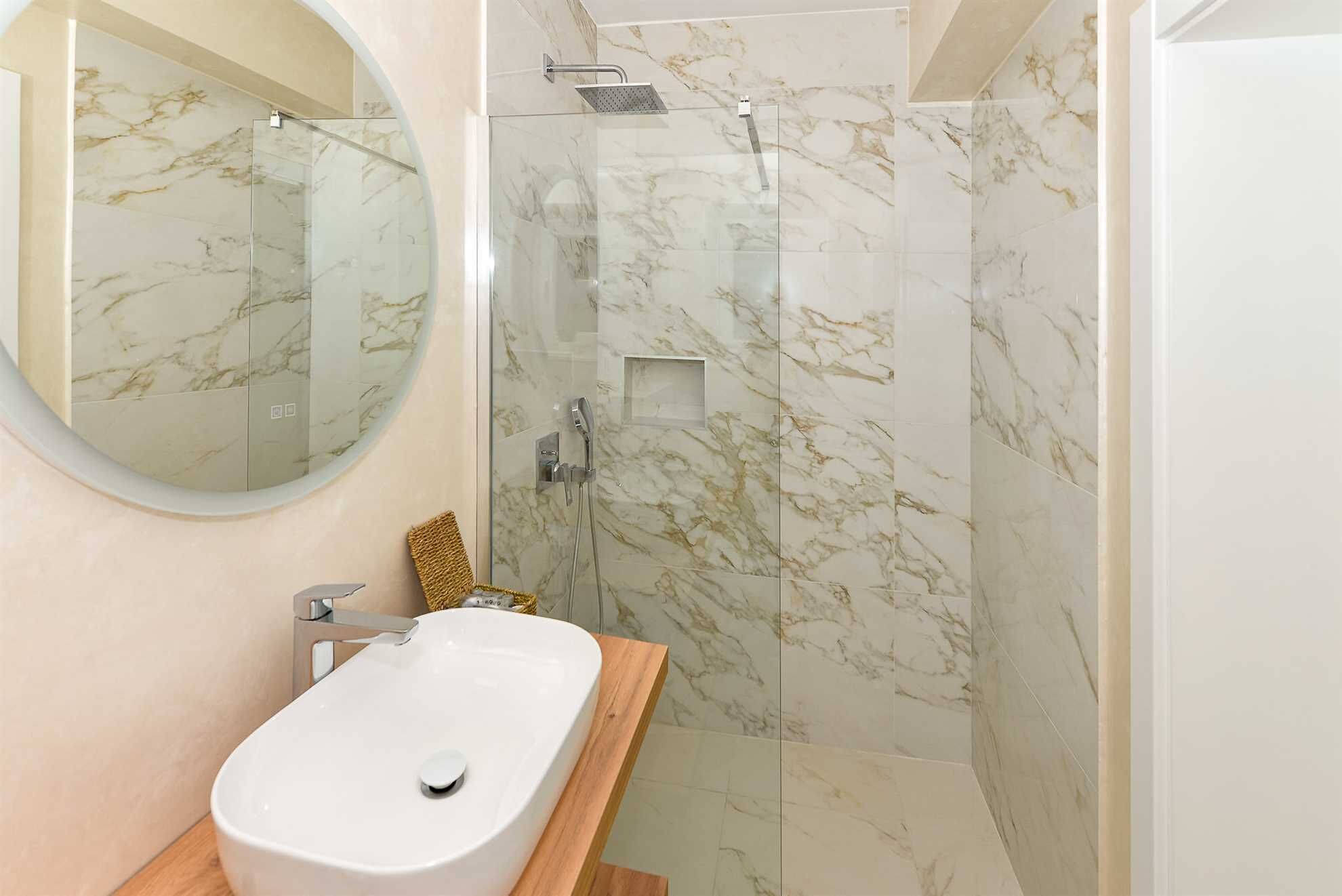 Un baño con ducha de mármol, lavabo blanco y espejo redondo.