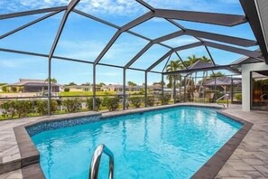 An indoor swimming pool with a glass ceiling, surrounded by a poolside area and a view of residential houses. - Nautical Dream Gulf Access Home (Cape Coral)