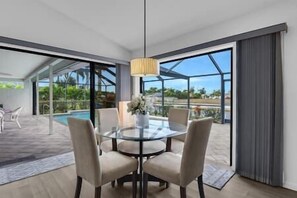A modern dining area with a glass table and chairs, a hanging light fixture, and large windows offering a view of a swimming pool and greenery. - Nautical Dream Gulf Access Home (Cape Coral)