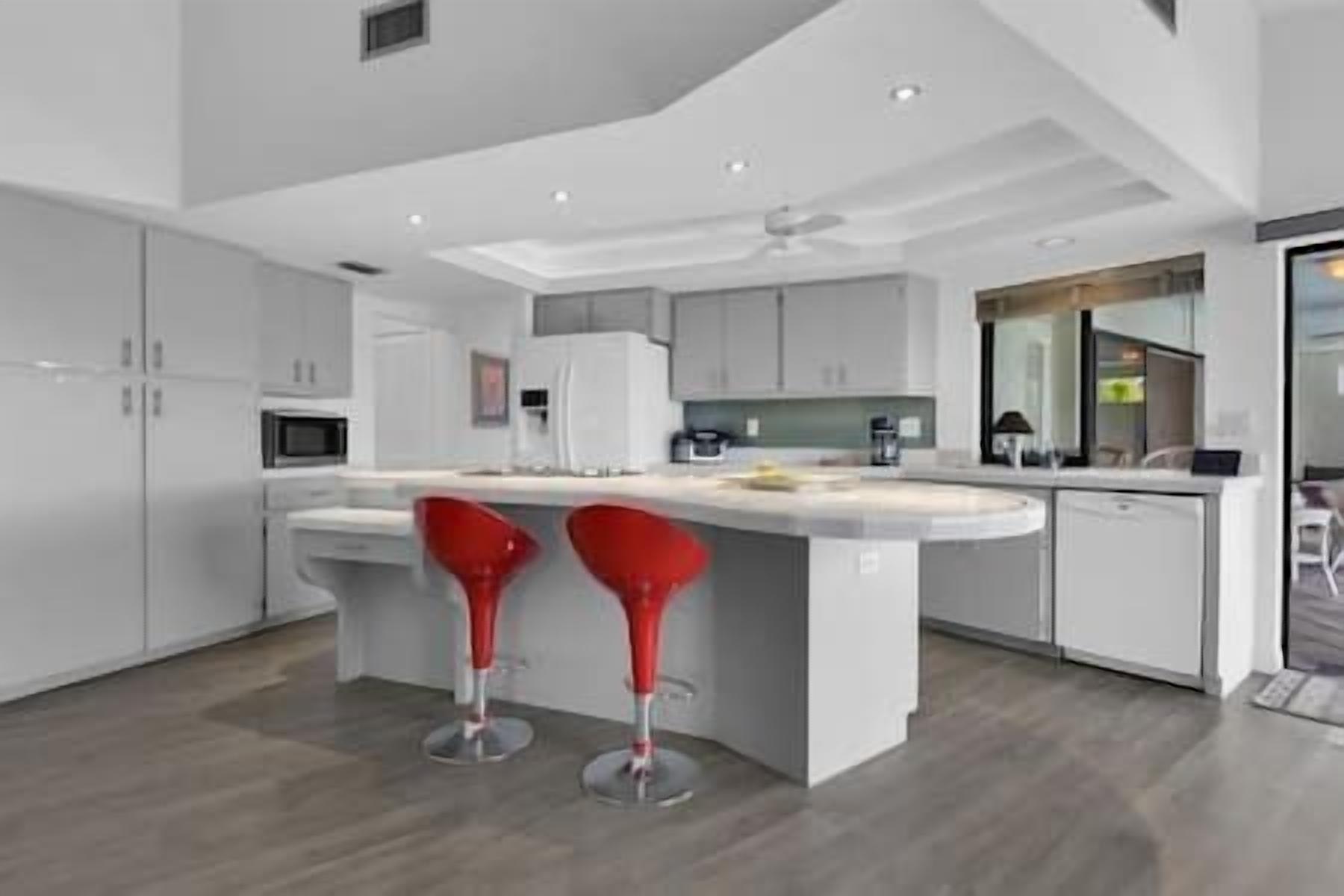A modern kitchen with a central island, two red bar stools, and a view into the adjacent living area.