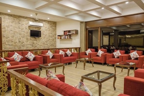Een ruime lobby met rode banken, houten bijzettafels, een stenen muur, een flatscreen-tv en een plafondventilator.