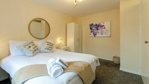 Una cama bien hecha con edredón blanco y manta color beige, un espejo redondo con marco dorado, una lámpara de noche y un cuadro en la pared.