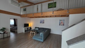 Amplia sala de estar con techo alto, vigas de madera, un sofá gris, una mesita y un televisor sobre un mueble blanco.