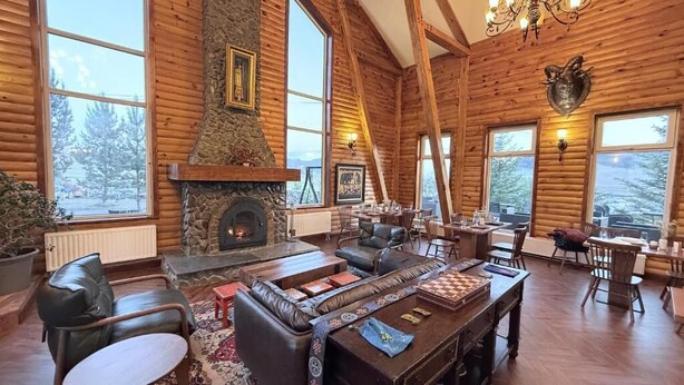 A log cabin interior with a stone fireplace, leather furniture, and large windows offering views of the surrounding forest. - Big Sky Lodge (Ulaanbaatar)