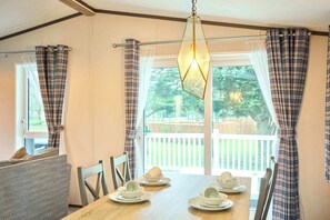 A dining area with a wooden table set for four, flanked by plaid curtains and a view of a deck and trees. - Host & Stay | POR01 Lotus Lodge (Pocklington)