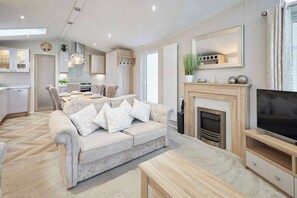 A modern living room with a sofa, coffee table, fireplace, TV, and kitchen area. - Host & Stay | The Braunston Lodge (Pocklington)