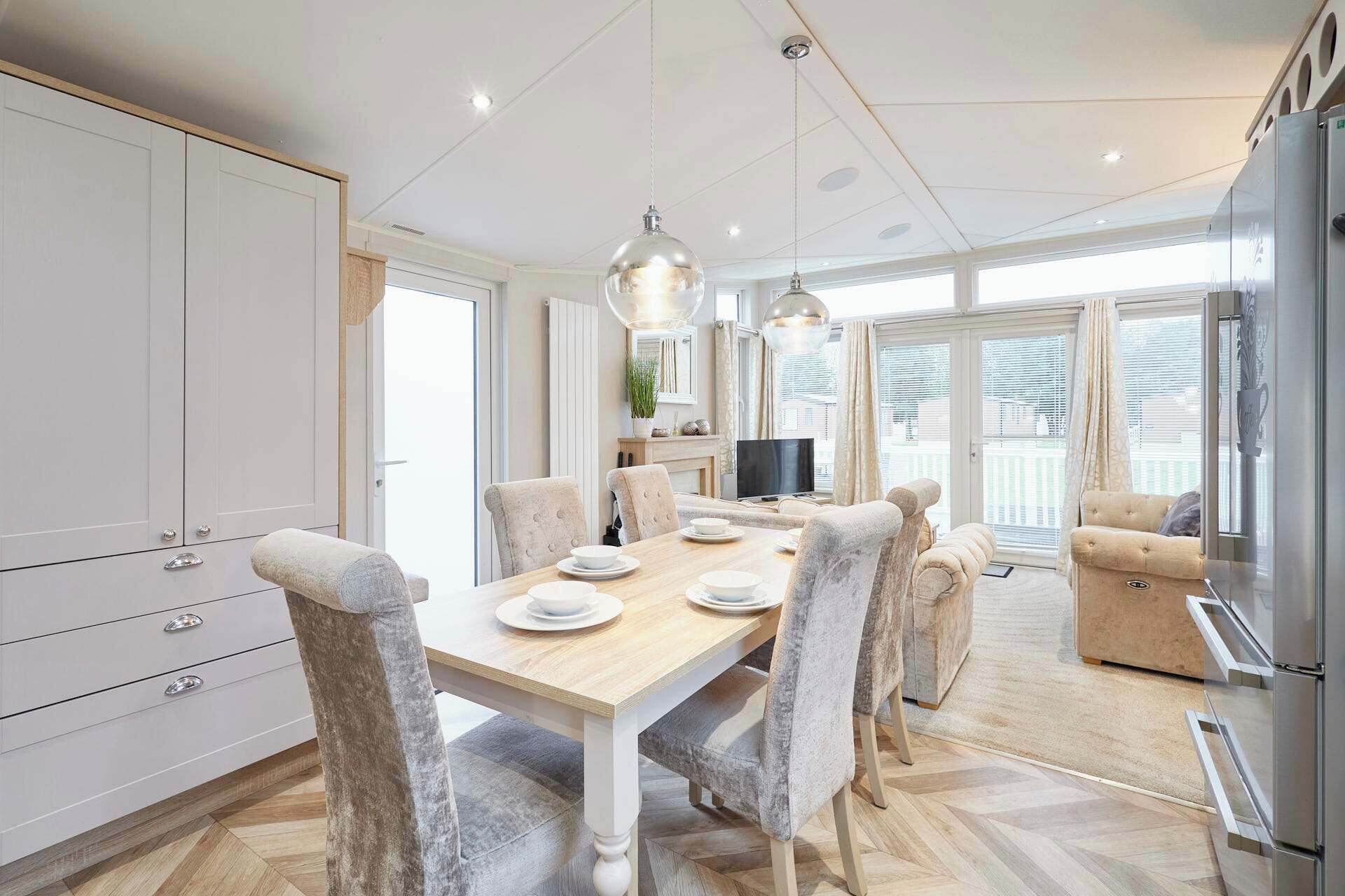A modern kitchen with a dining area, featuring a wooden table, grey upholstered chairs, pendant lighting, and a built-in TV.