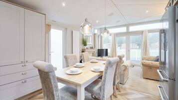 A modern kitchen with a dining area, featuring a wooden table, grey upholstered chairs, pendant lighting, and a built-in TV.