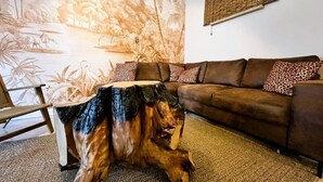 Un soggiorno con un divano marrone, cuscini a stampa leopardo, un tavolino in legno e un murale raffigurante una scena tropicale.