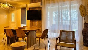 Un'area pranzo con un tavolo rotondo in legno, sei sedie, una TV a schermo piatto montata a parete e un'ampia finestra con tende leggere.
