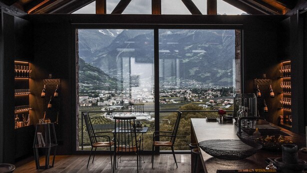 A modern interior with a large window offering a mountain view, a bar area with wine glasses, and a seating arrangement. - Pratenberg Apartment Weite Penthouse with Mountain View, Sauna & Saltwater Rooftop Pool (Merano)