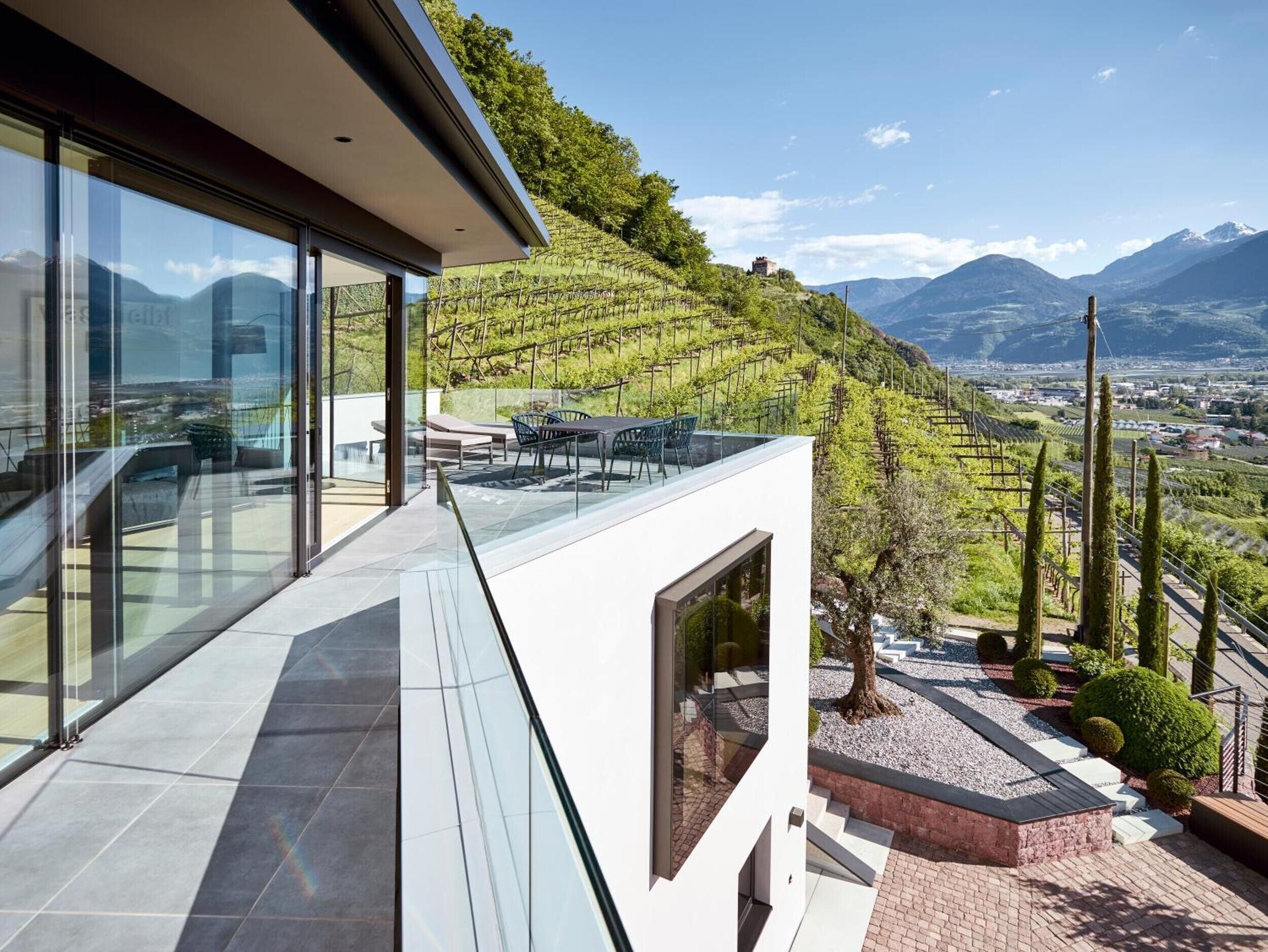 A modern building with large glass windows, a terrace with outdoor furniture, and a vineyard view.