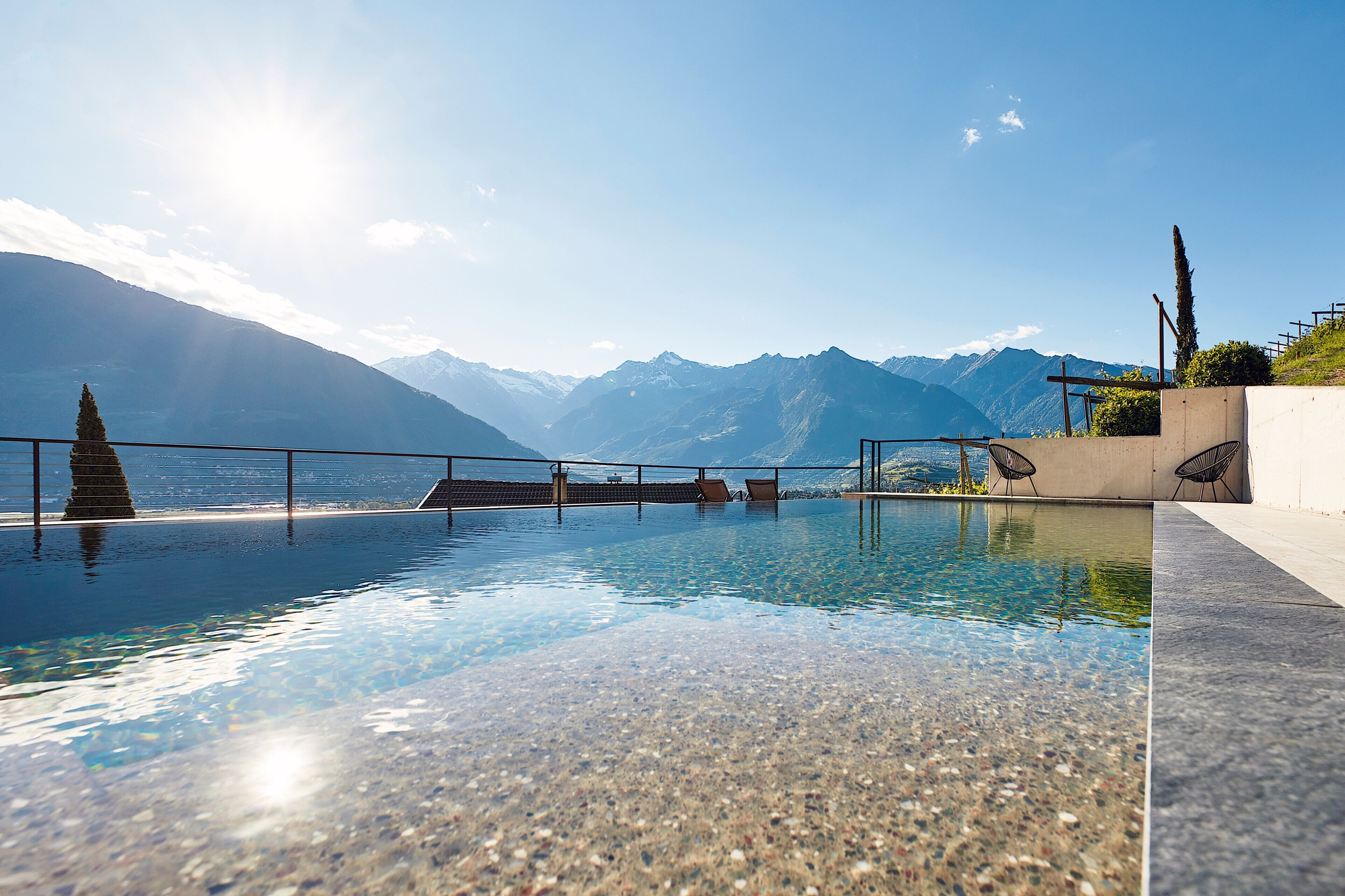 An infinity pool with clear water, a mountain backdrop, and a few lounge chairs.