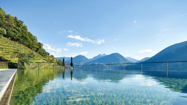 Una piscina cristalina con vista a la montaña, una terraza de madera y una barandilla de metal.