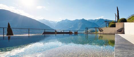 An infinity pool with clear water, a mountain backdrop, and a few lounge chairs.