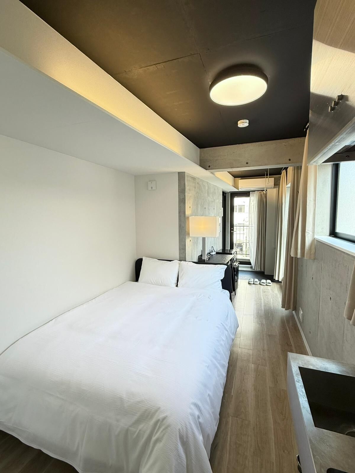 A modern hotel room with a large bed, a desk, a chair, a lamp, and a view through a window.