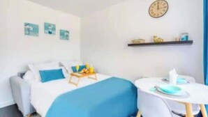 Una camera d'albergo con un letto, un tavolino con una sedia, una sveglia appesa al muro e uno scaffale con vari oggetti.