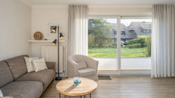 A living room with a sofa, armchair, and round coffee table. There is a radiator, a floor lamp, and a view of a garden with a thatched-roof house. - Owl's Nest - Wenningstedt-Braderup (Sylt) (Wenningstedt-Braderup (Sylt))