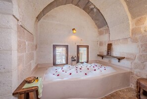 A stone-walled room with a large bathtub filled with water and rose petals, two windows with blinds, and a small table with a towel and bottles. - Tol Of Cappadocia (Nevsehir)