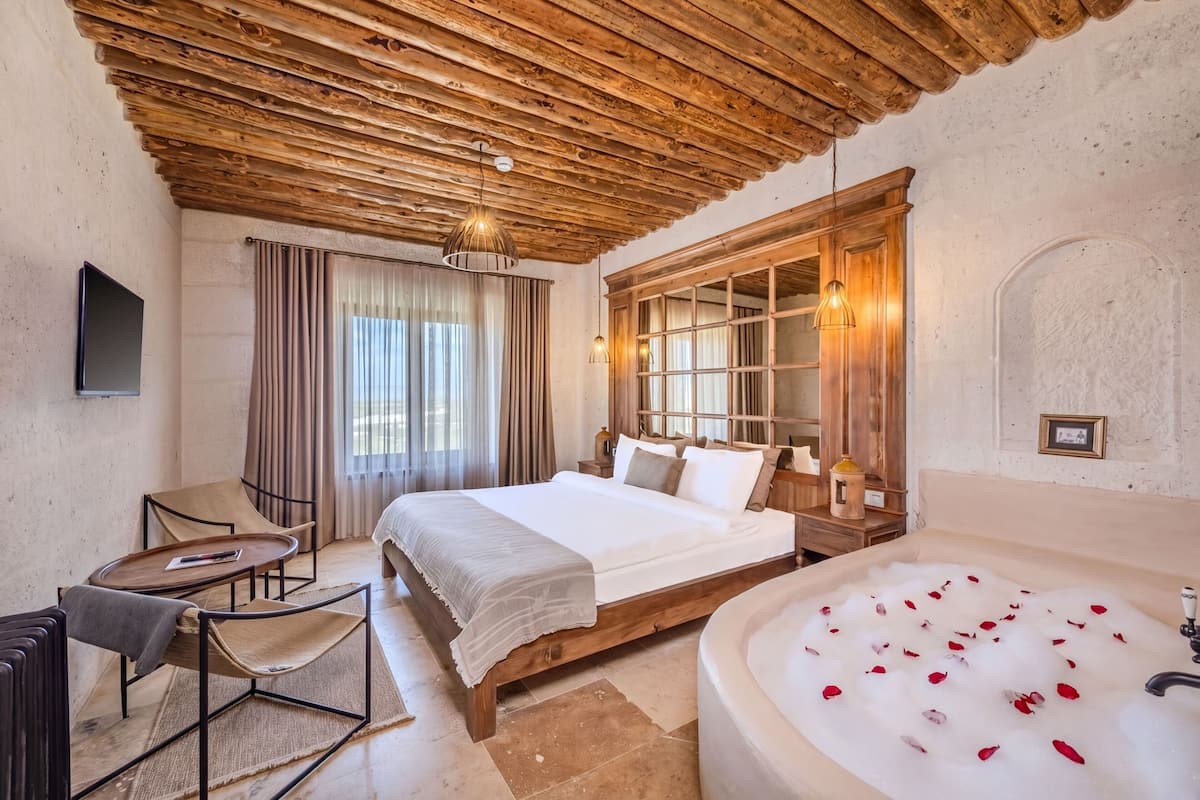 A bedroom with a wooden ceiling, a large bed, a sitting area with a table and chairs, a bathtub with rose petals, and a window with curtains.