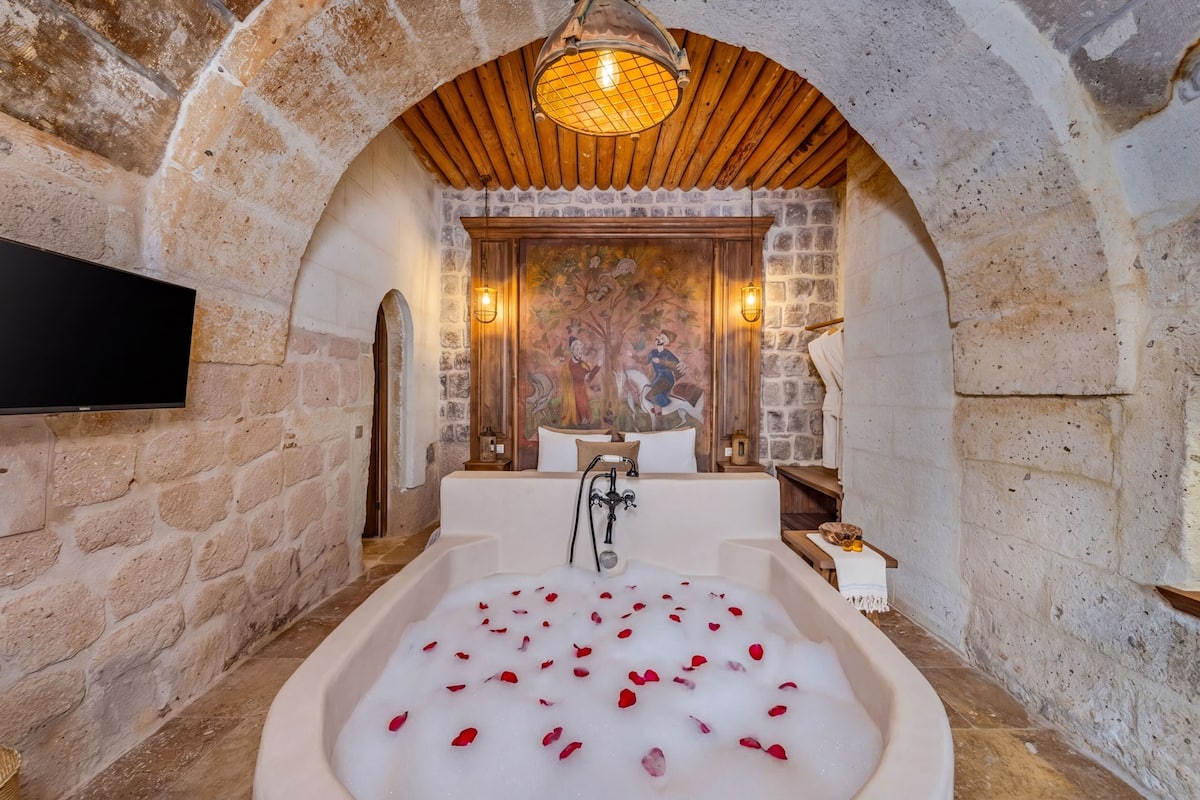 A stone-walled bathroom with a bathtub filled with rose petals, a wooden ceiling, and a wall-mounted TV.