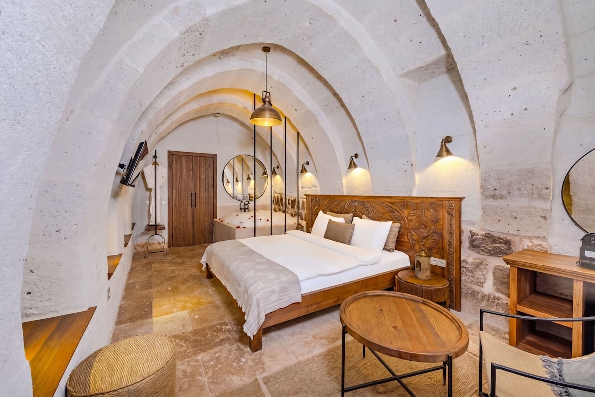 A bedroom with a wooden bed, a round wooden table, a wicker chair, and a stone wall.