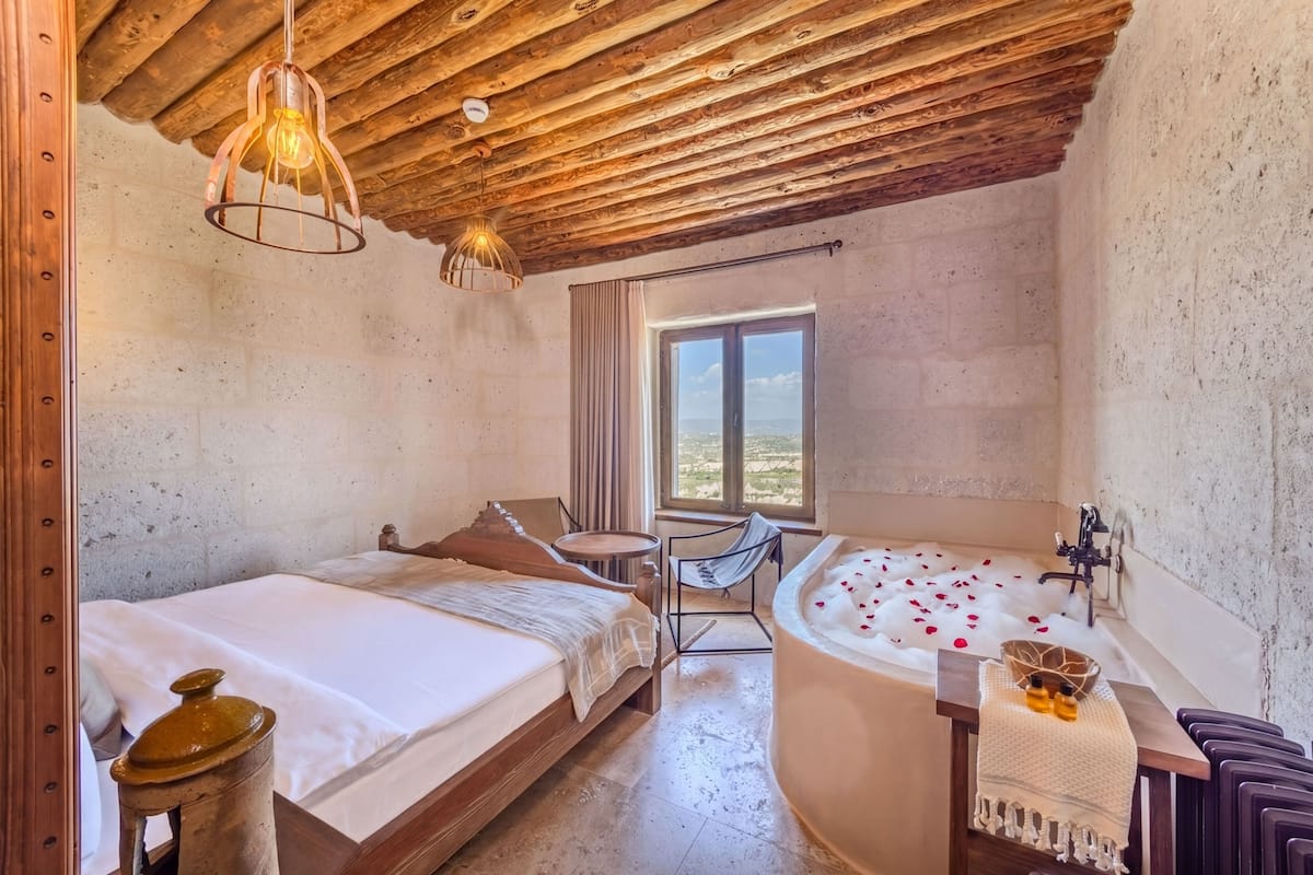 A room with a wooden ceiling, a bed, a chair, a bathtub with rose petals, and a window with a view.