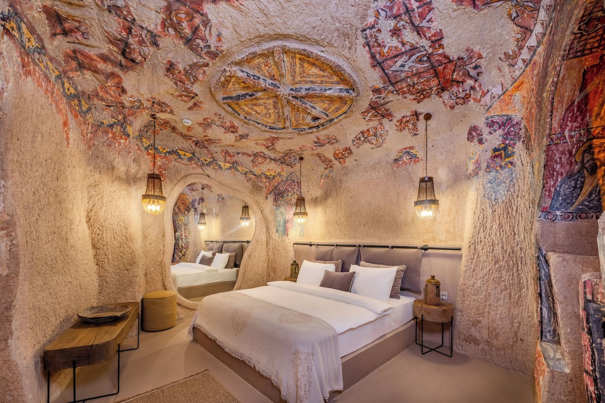 A room with a large bed, a wooden bench, a small table, and a wall-mounted lamp.