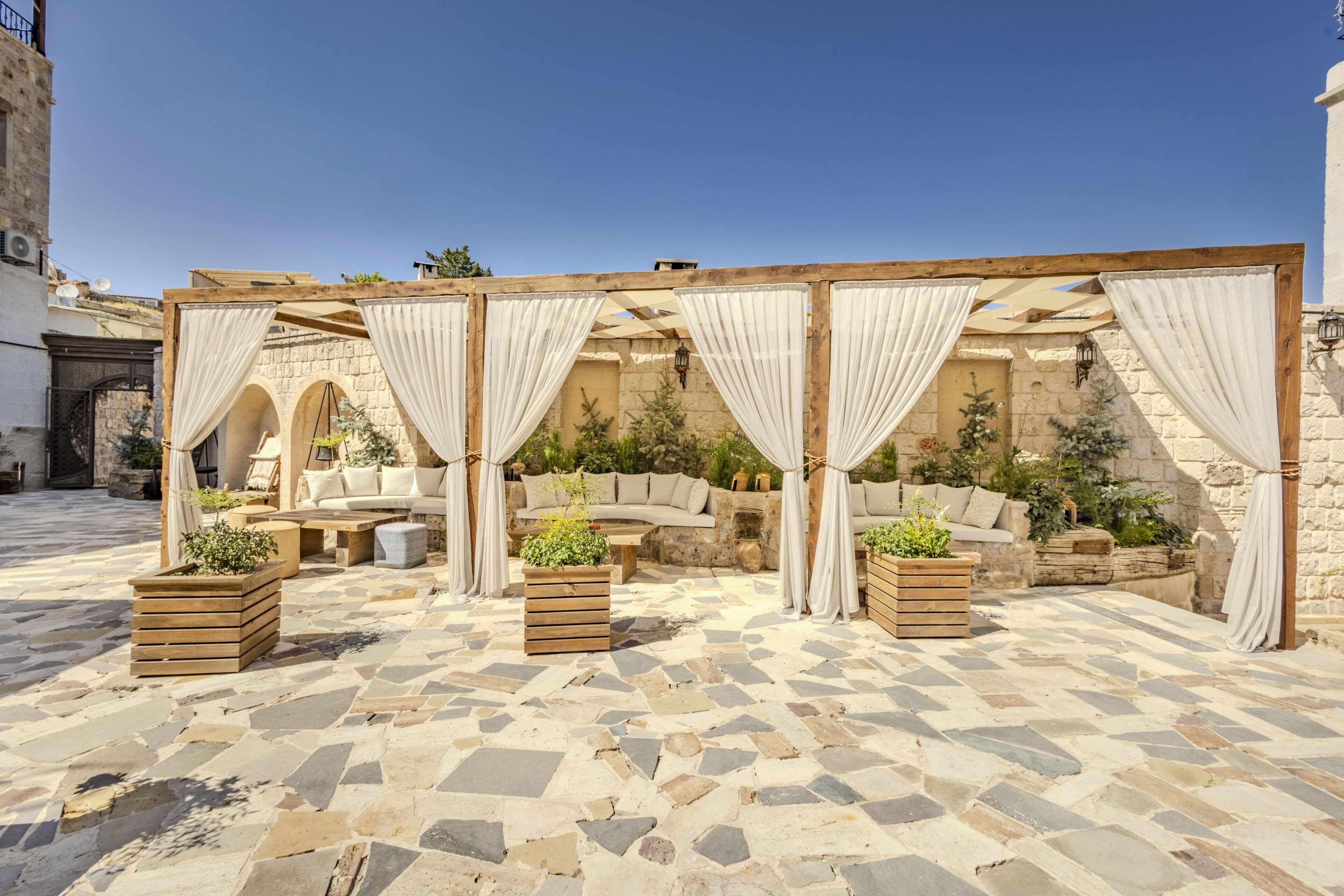 Un espacio de estar al aire libre con suelo de piedra, muebles de madera y cortinas blancas.