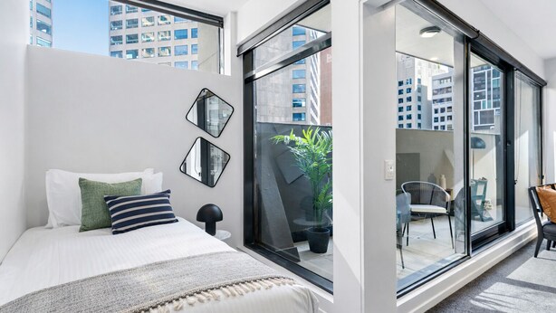 Ein modernes Hotelzimmer mit einem großen Bett, einem kleinen Tisch, einem Sessel und einer Schiebetür zu einem Außenbereich mit Blick auf die Stadt.