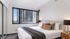 Ein modernes Schlafzimmer mit großem Fenster, einem Bett mit vielen Kissen, einem Nachttisch mit Lampe und Blick auf hohe Gebäude.