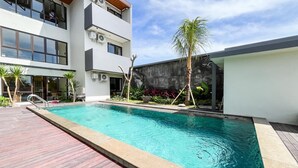 Una piscina con bordo in legno, un edificio moderno con ampie finestre e una palma.
