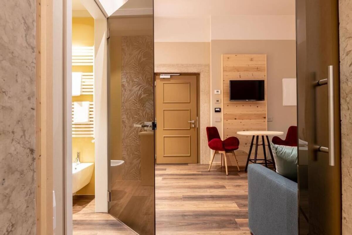 A modern hotel room with a wooden floor, a sofa, a TV mounted on the wall, and a door leading to a bathroom.