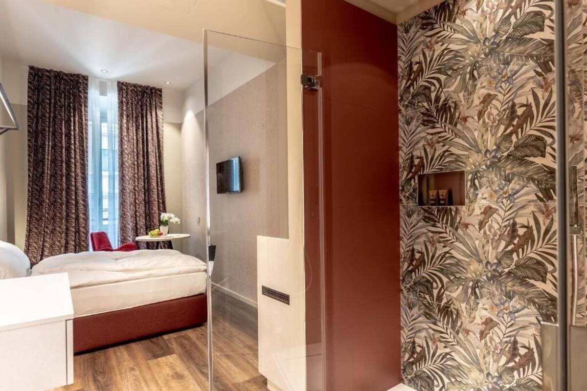 A modern hotel room with a bed, a glass partition, a small table with a vase, and a television.