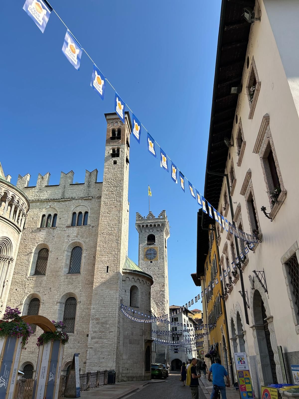 Un bâtiment historique en pierre, doté d’une tour, une rue décorée de bannières et un ciel d’un bleu éclatant.