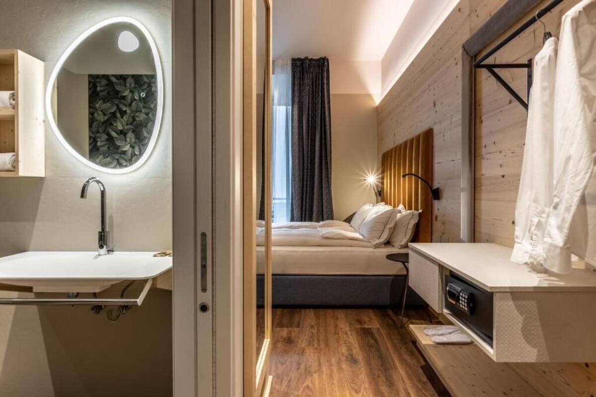 A modern bedroom with a wooden floor, a bed with white bedding, a bedside table with a remote control, a built-in circular mirror, and a shower area with a curtain.