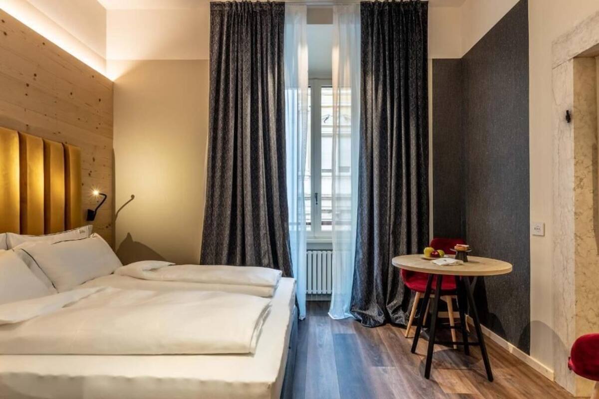 A hotel room with a bed, a small table with a chair, and a window with curtains.