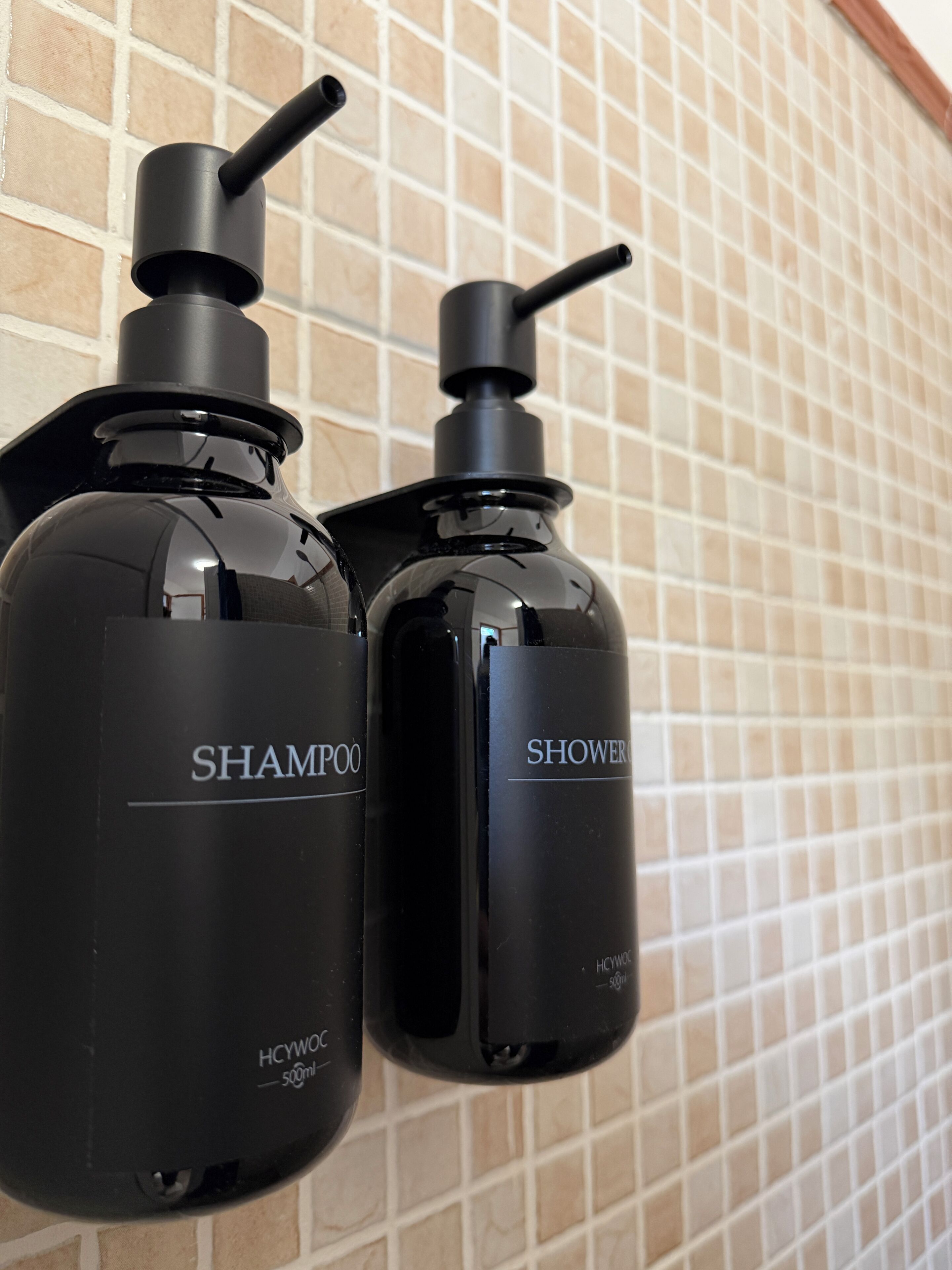 Zwei schwarze Flaschen mit Shampoo und Duschgel vor einer gefliesten Wand.