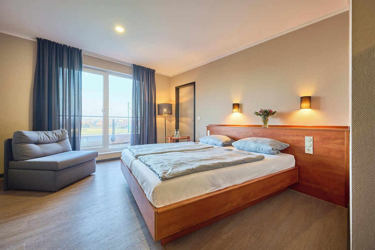A hotel room with a large bed, a sofa, a nightstand, a lamp, and a window with curtains.
