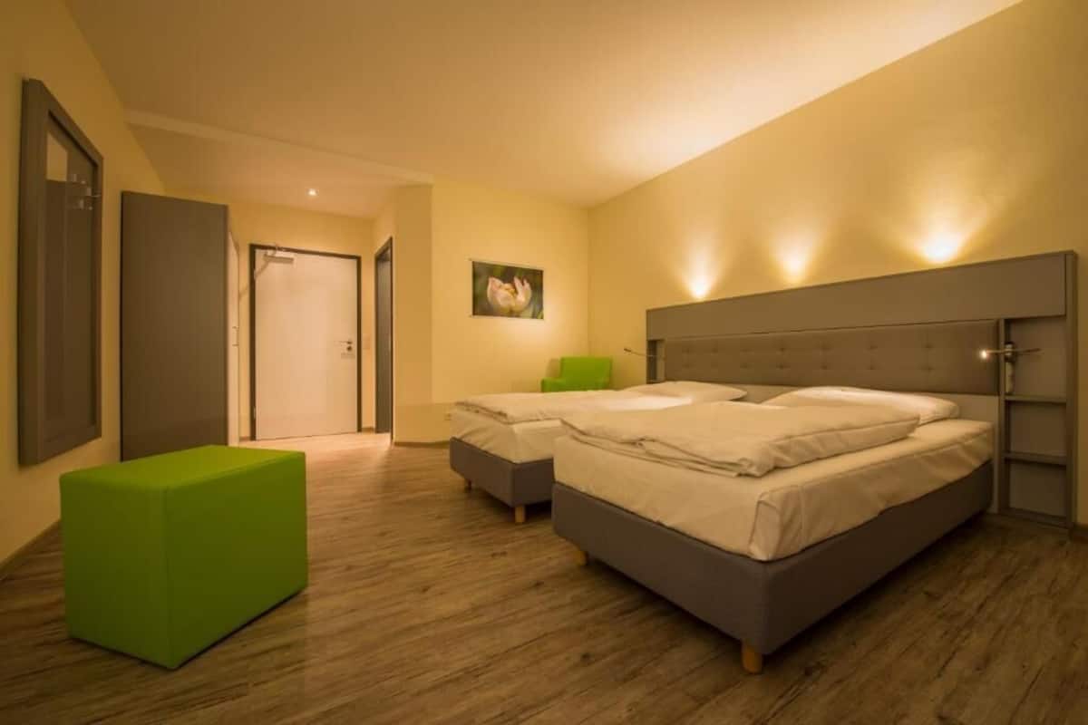 A bedroom with two beds, a green ottoman, and a painting on the wall.