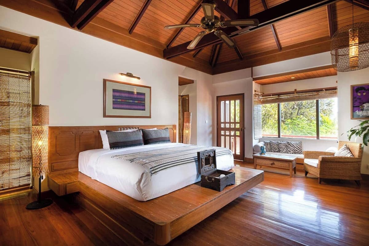 Un dormitorio amplio con una cama grande, piso de madera, ventilador de techo y vistas a zonas verdes a través de amplias ventanas.