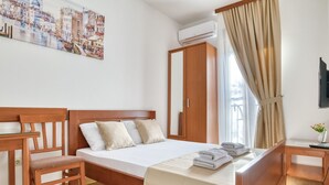 Hotellihuone, jossa on puinen sänky, työpöytä, tuoli, suuri ikkuna verhoilla, ilmastointilaite ja kaupunkimaisemaa esittävä taulu.