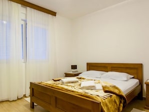 A hotel room with a wooden bed, white linens, a nightstand, and a window with sheer curtains.