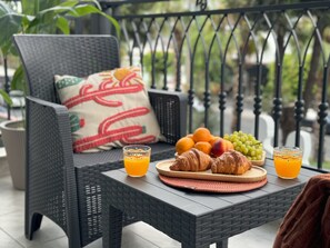 Uma cadeira de vime com almofada, uma mesa com comida e bebida, e um corrimão ao fundo.