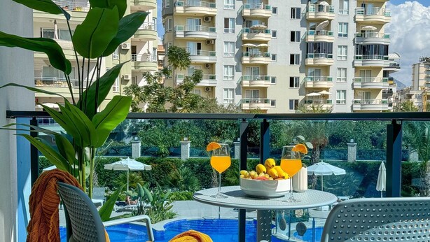 A balcony with a pool, chairs, a table with fruit and drinks, and a view of a multi-story building. - Qoople Aria-07 neue Wohnung im Stadtzentrum by Interhome (Alanya)