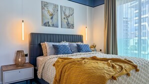 Een moderne slaapkamer met een groot bed, twee aan de muur hangende schilderijen, een bedlampje en uitzicht op de stad door dunne gordijnen.
